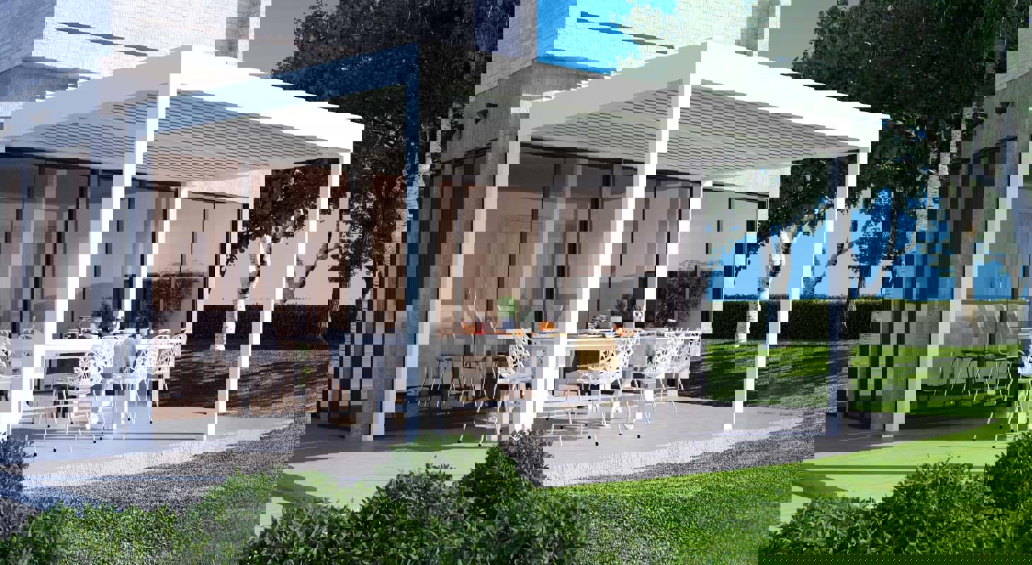 Sodobna bela pergola ob hiši z veliko steklenimi površinami, pod katero je jedilna garnitura na terasi, obdana z zelenico in drevesi.