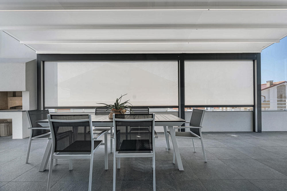 Moderna terasa s pergolo in vgrajenimi screen roloji za zaščito pred soncem, opremljena z jedilno garnituro v črno-beli barvni kombinaciji.