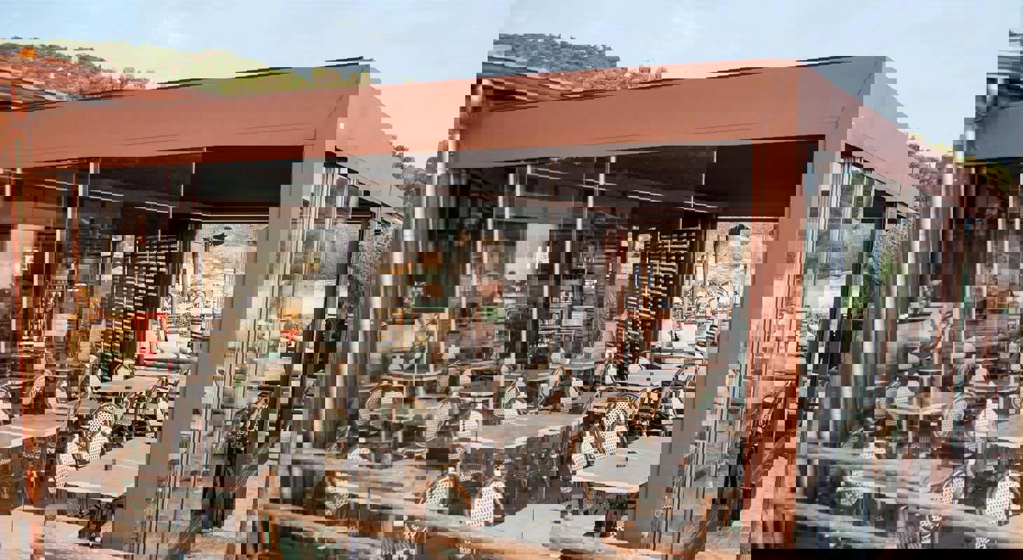 Zunanja terasa restavracije z moderno rjavo pergolo in steklenimi stenami, opremljena z elegantnimi mizami in stoli ob plaži.