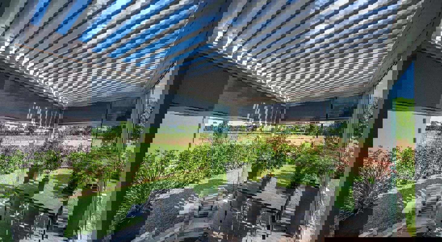 Moderna bioklimatska pergola z lamelami, senčili in steklenimi stenami na vrtni terasi s pogledom na naravo.