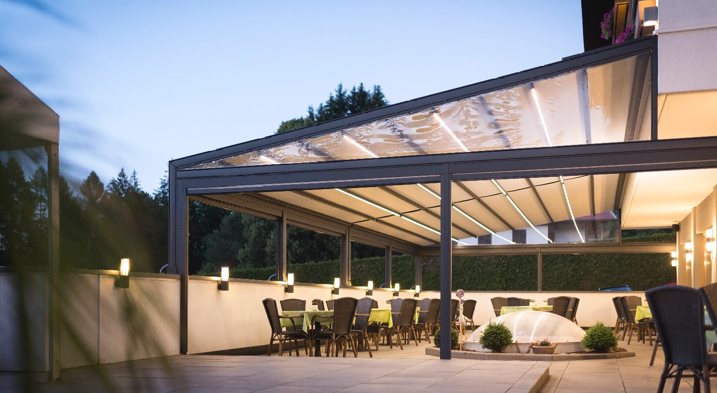 Velika pergola z LED osvetlitvijo in prozorno streho, postavljena nad zunanjo restavracijsko teraso z več mizami in stoli v večernem ambientu.