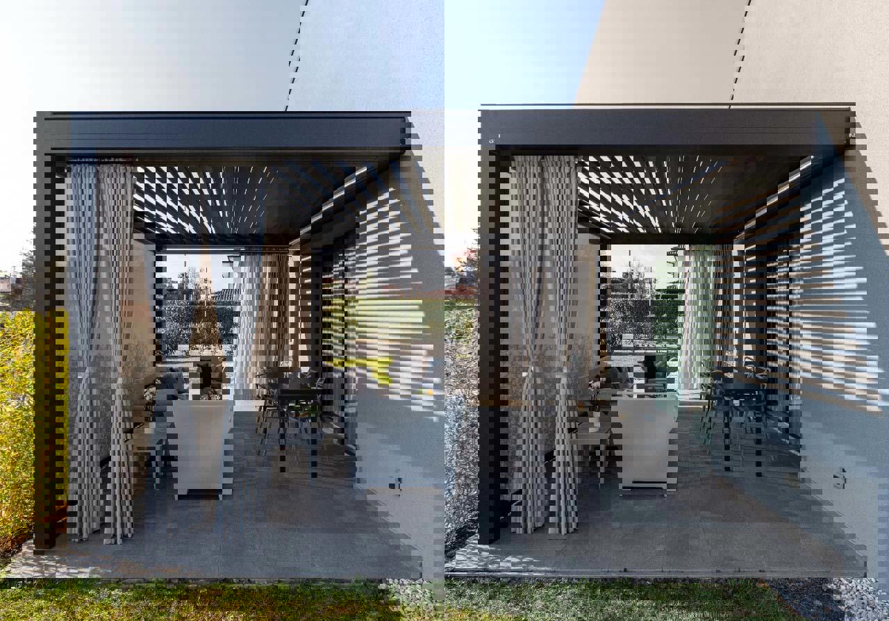 Sodobna bioklimatska pergola s črnimi lamelami in zavesami ob hiši, opremljena z vrtno sedežno garnituro in jedilno mizo na tlakovanem dvorišču.