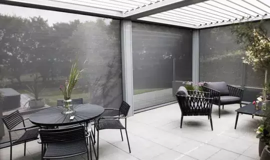 Moderna pergola z vrtljivimi lamelami in stranskimi screen roloji, opremljena z vrtnim pohištvom na tlakovanem dvorišču, obdana z zelenjem.