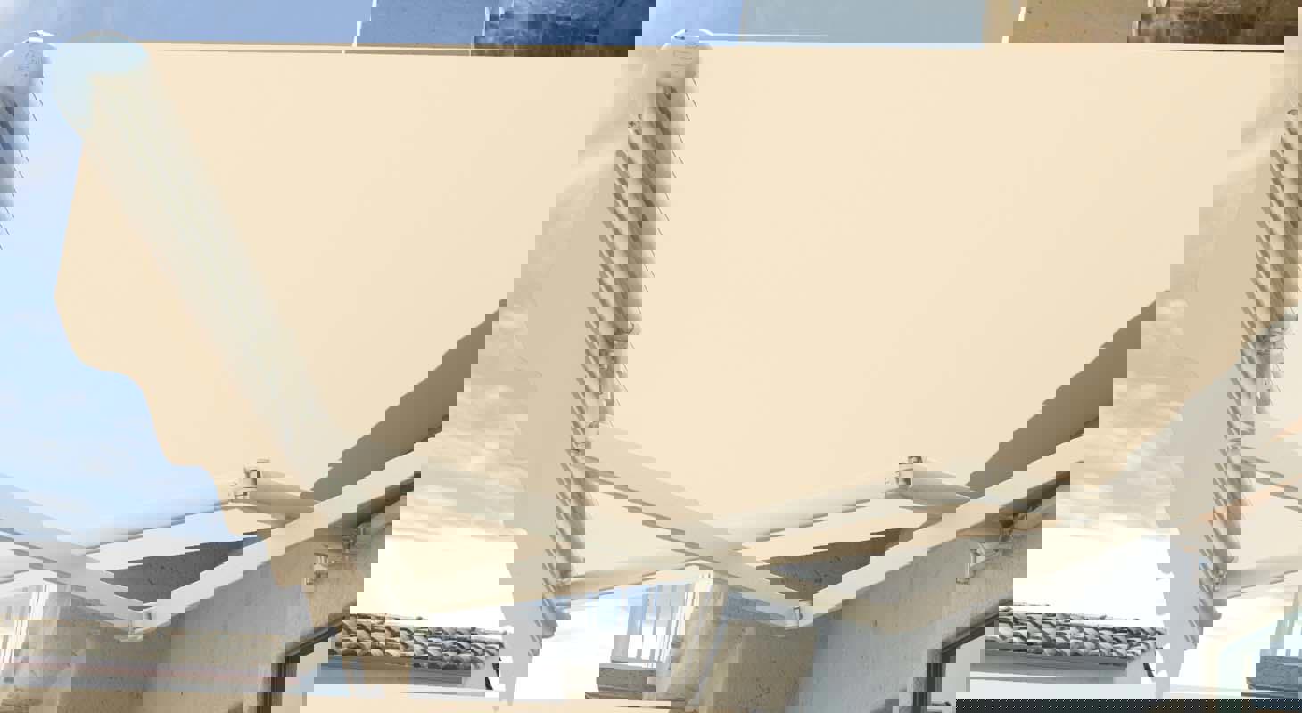 Bež tenda z valovitim robom in zložljivim mehanizmom, pritrjena na betonsko fasado, pogled od spodaj navzgor.