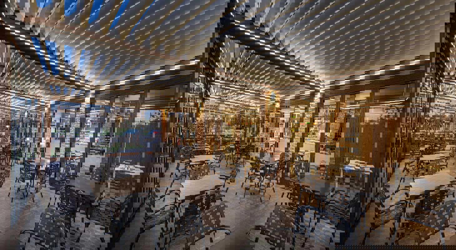 Zunanja pokrita terasa restavracije z bioklimatsko pergolo in ambientno LED osvetlitvijo ter črnimi kovinskimi stoli in mizami.