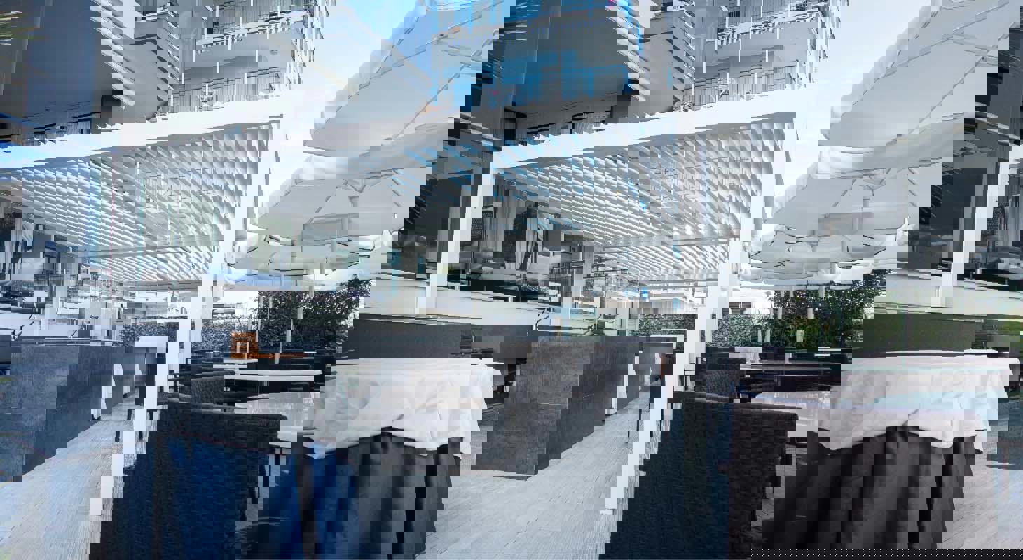 Bela bioklimatska pergola s premičnimi lamelami ob sodobni restavraciji na terasi hotela, obdana z jedilnimi mizami, stoli in senčniki.