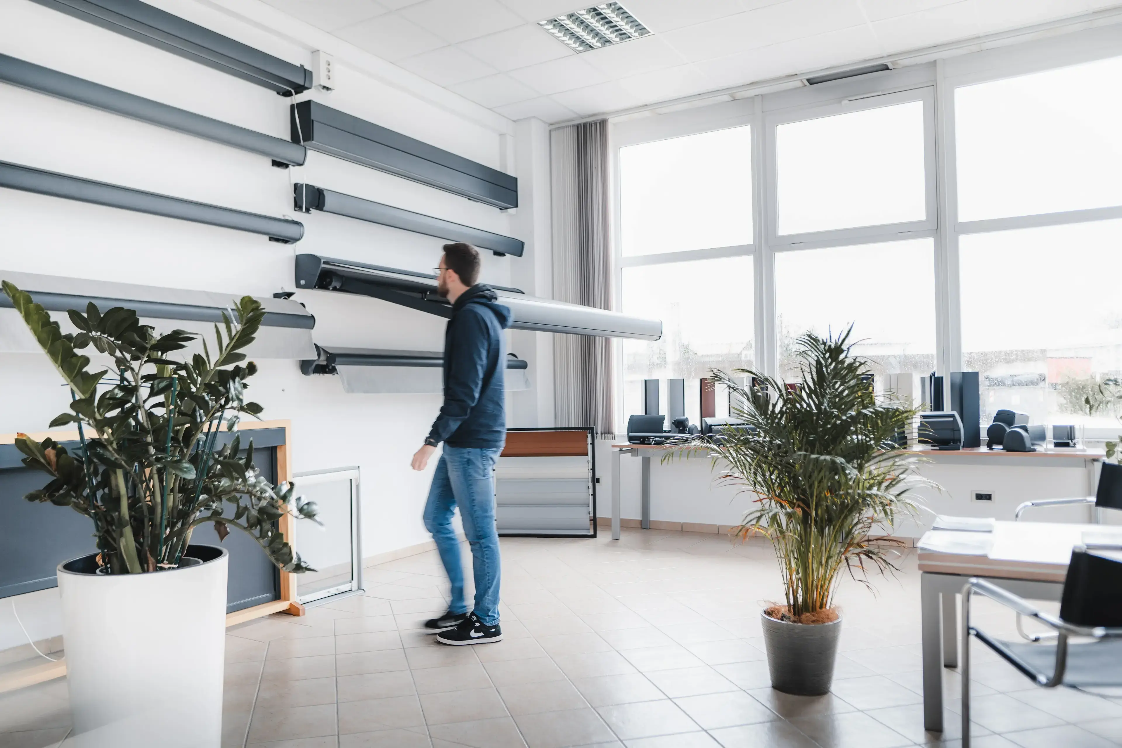 Moški v razstavnem salonu z več modeli tend na steni, v svetlem prostoru z velikimi okni in okrasnimi rastlinami.