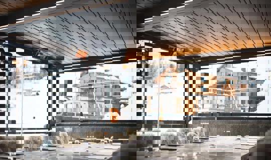 Moderna zaprta terasa s kovinsko streho in steklenimi stenami, opremljena z ogrevalnimi paneli in udobnim pohištvom, s pogledom na zasneženo gorsko vas in leseno kočo.