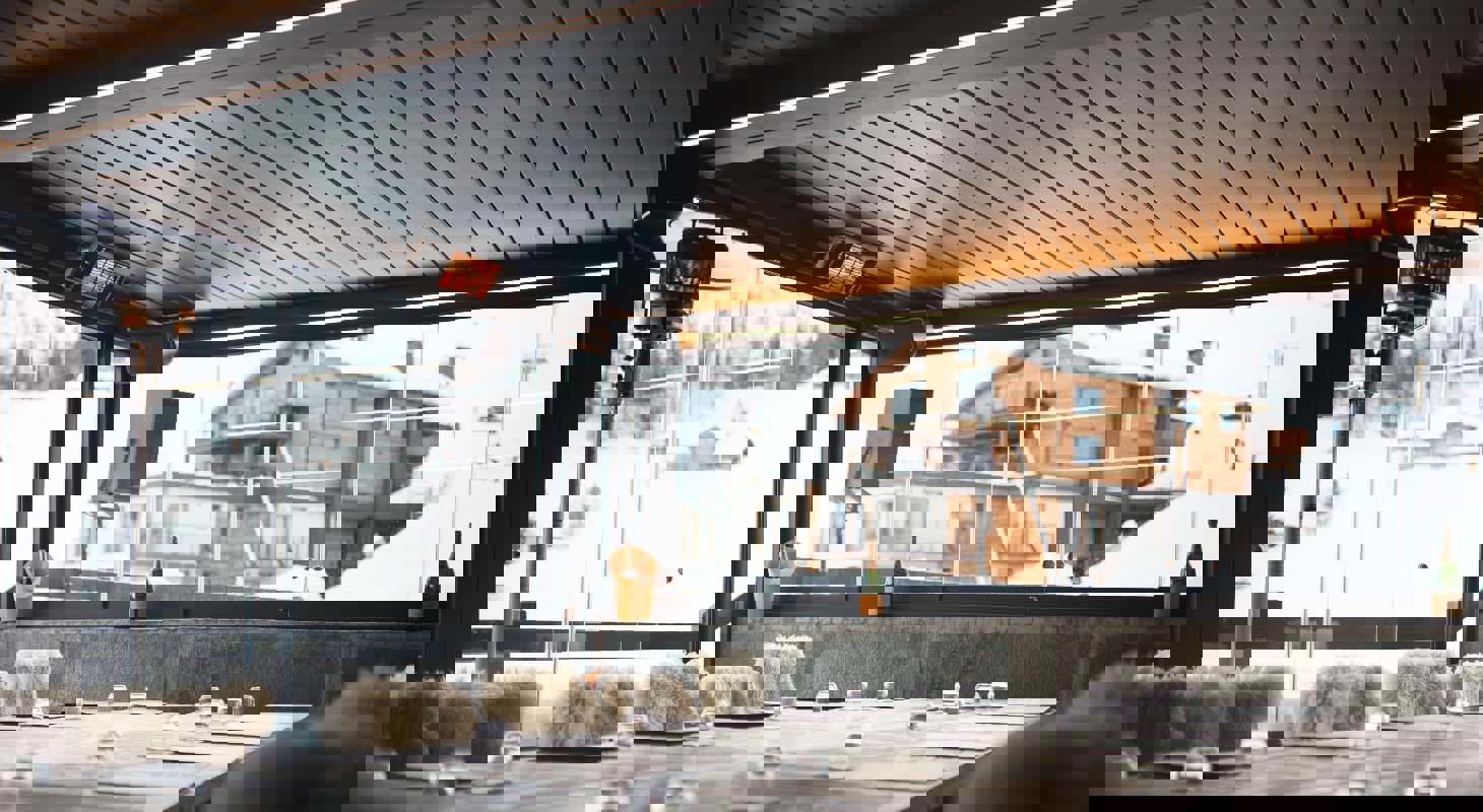 Moderna zaprta terasa s kovinsko streho in steklenimi stenami, opremljena z ogrevalnimi paneli in udobnim pohištvom, s pogledom na zasneženo gorsko vas in leseno kočo.